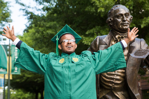 Graduate dressed in George Mason University graduation regalia lifts arms in jubilation as they pose with the bronze statue of George Mason 