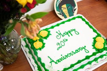 A cake with white, green, and yellow frosting says Happy Anniversary. A Mason Patriot image sits beside it on the table, with a vase of flowers
